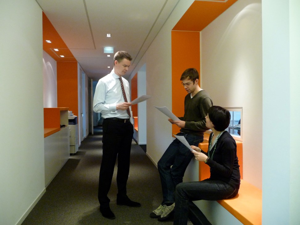 Modern office corridor with orange architectural elements and three professionals reviewing documents.