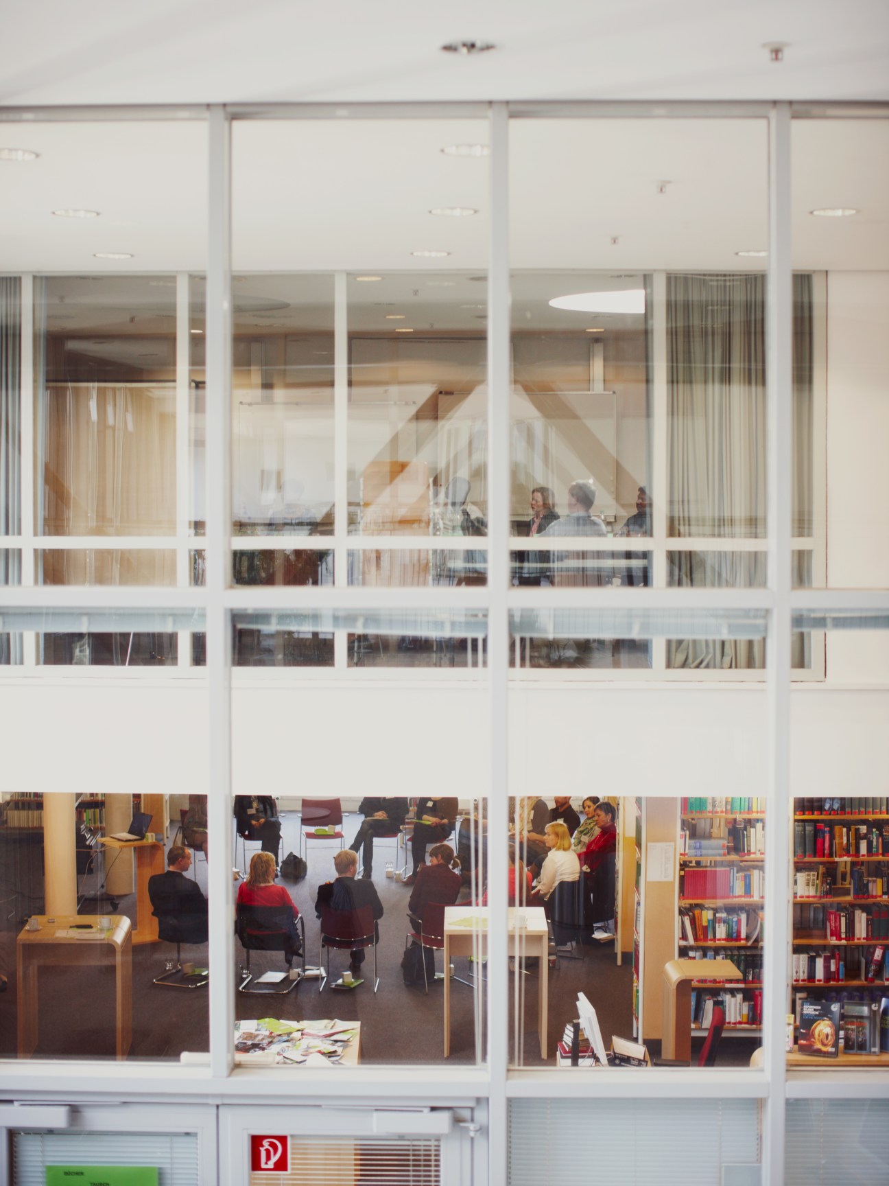 A conference taking place inside the Goethe-Institut Munich headquarters, visible through large glass windows with participants seated indoors.