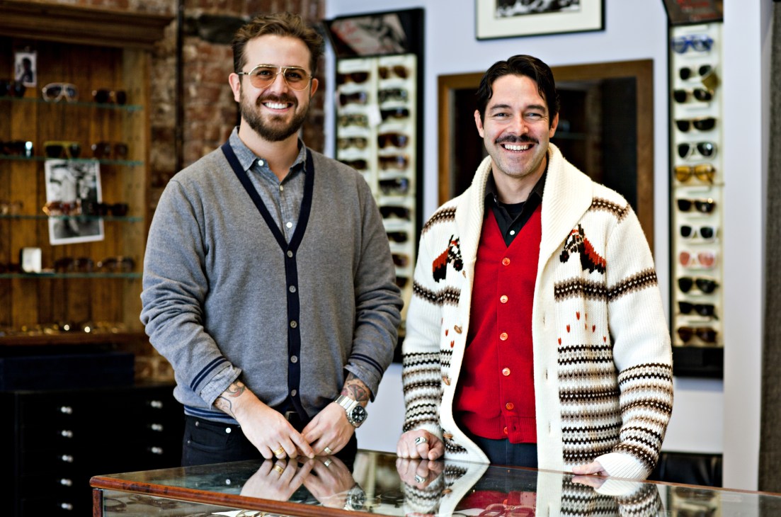 Jordan Silver and Erik Sacher stand at counter in their eyewear boutique with vintage frames displayed on walls behind them.