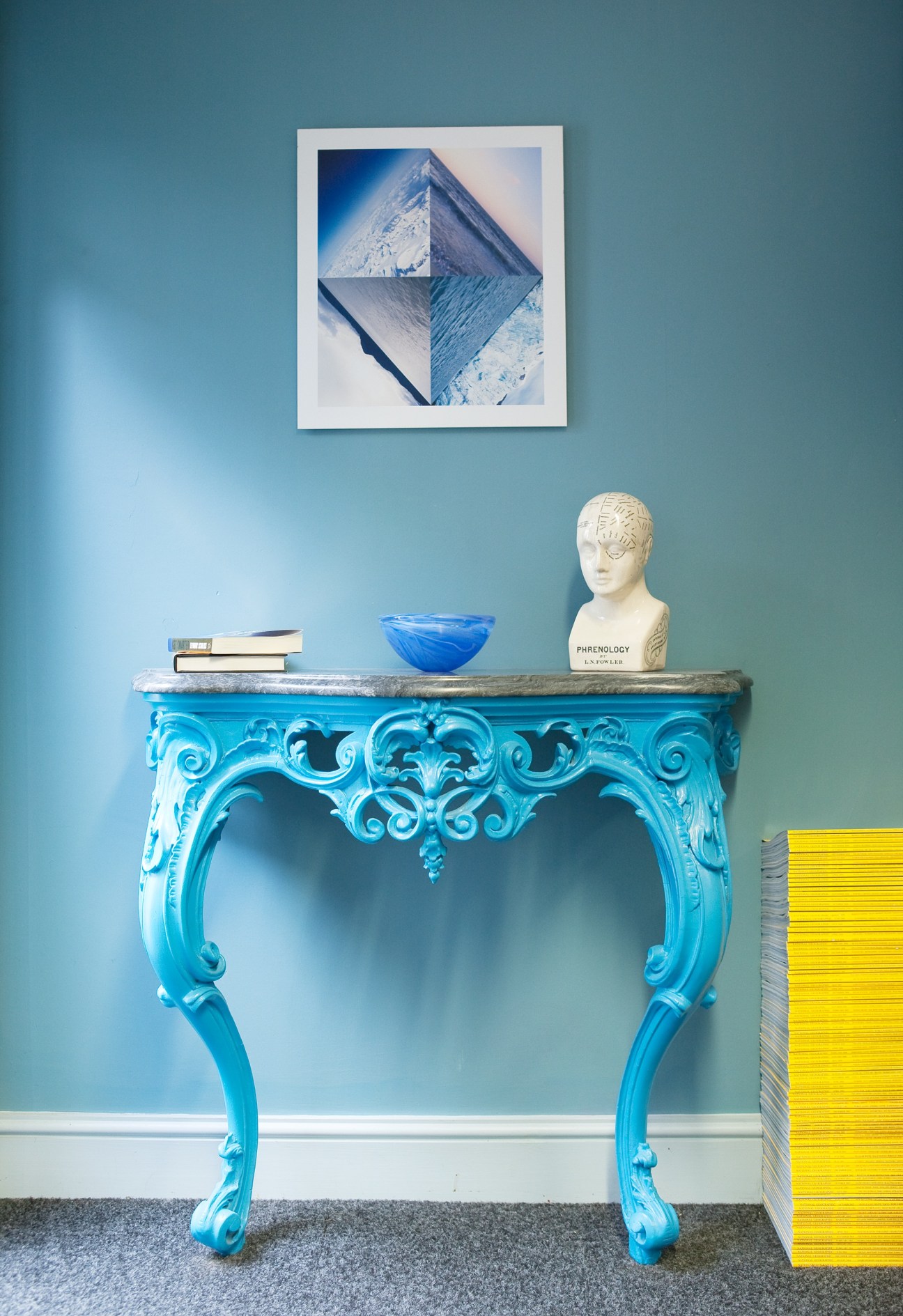 Bright blue ornate console table against teal wall with stacked books, blue bowl, and white bust sculpture.
