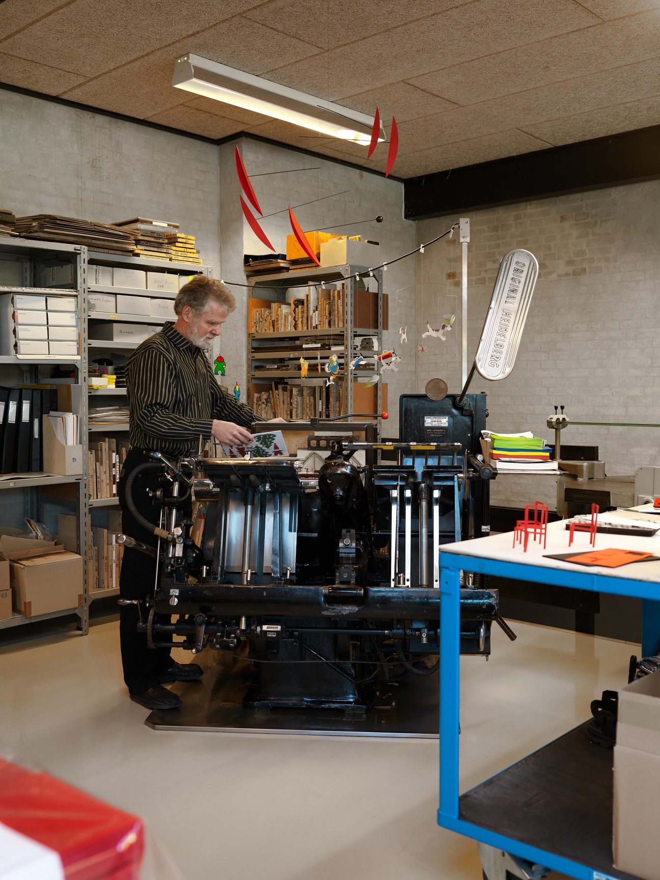 Ole Flensted operates a Heidelberg printing press in his workshop, with colorful mobiles hanging above.