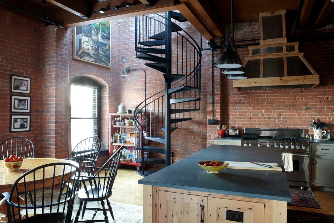 Modern loft kitchen with exposed red brick walls, spiral staircase, wooden beams, and island counter.