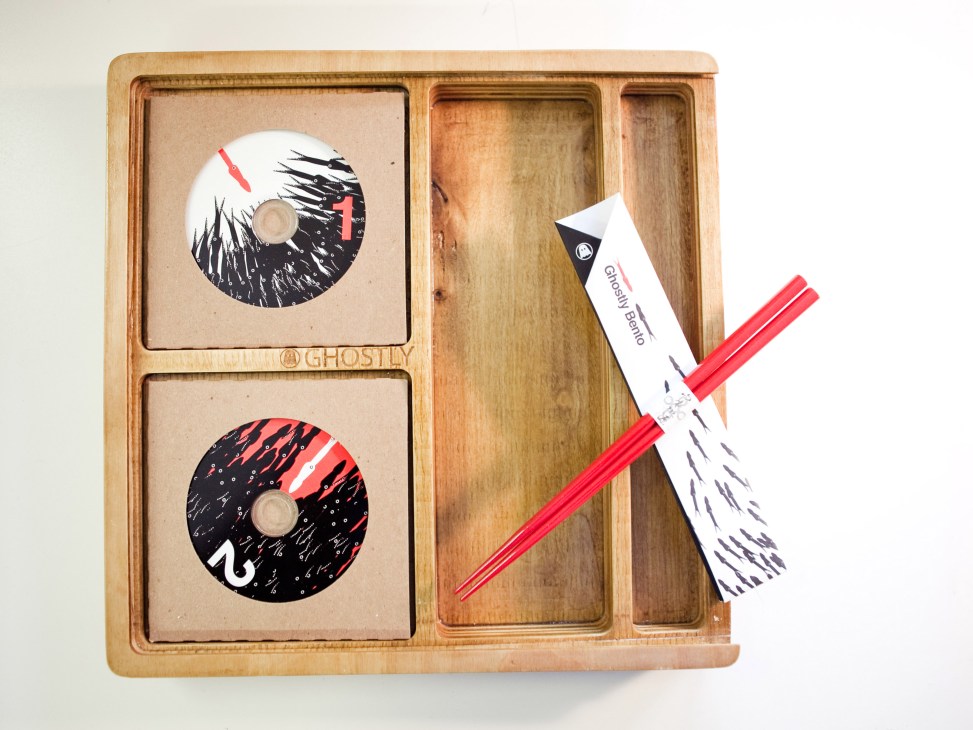 Wooden bento box containing two CDs with artistic black and red designs, chopsticks, and a Ghostly Berlin card.