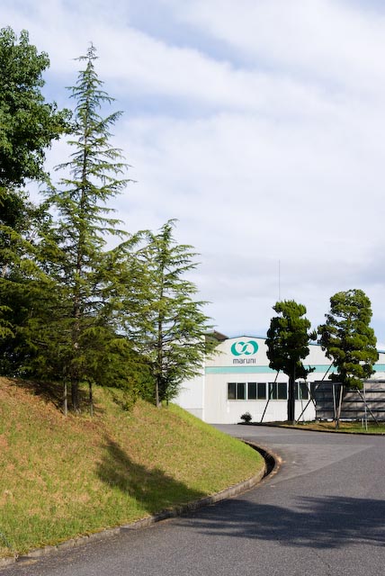 Maruni headquarters building with green logo visible on white facade, surrounded by wooded hills and landscaping.