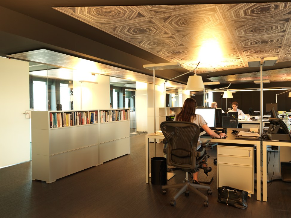 IJ Partners Geneva office with staff working at desks under decorative ceiling lights and white storage with bookshelves.