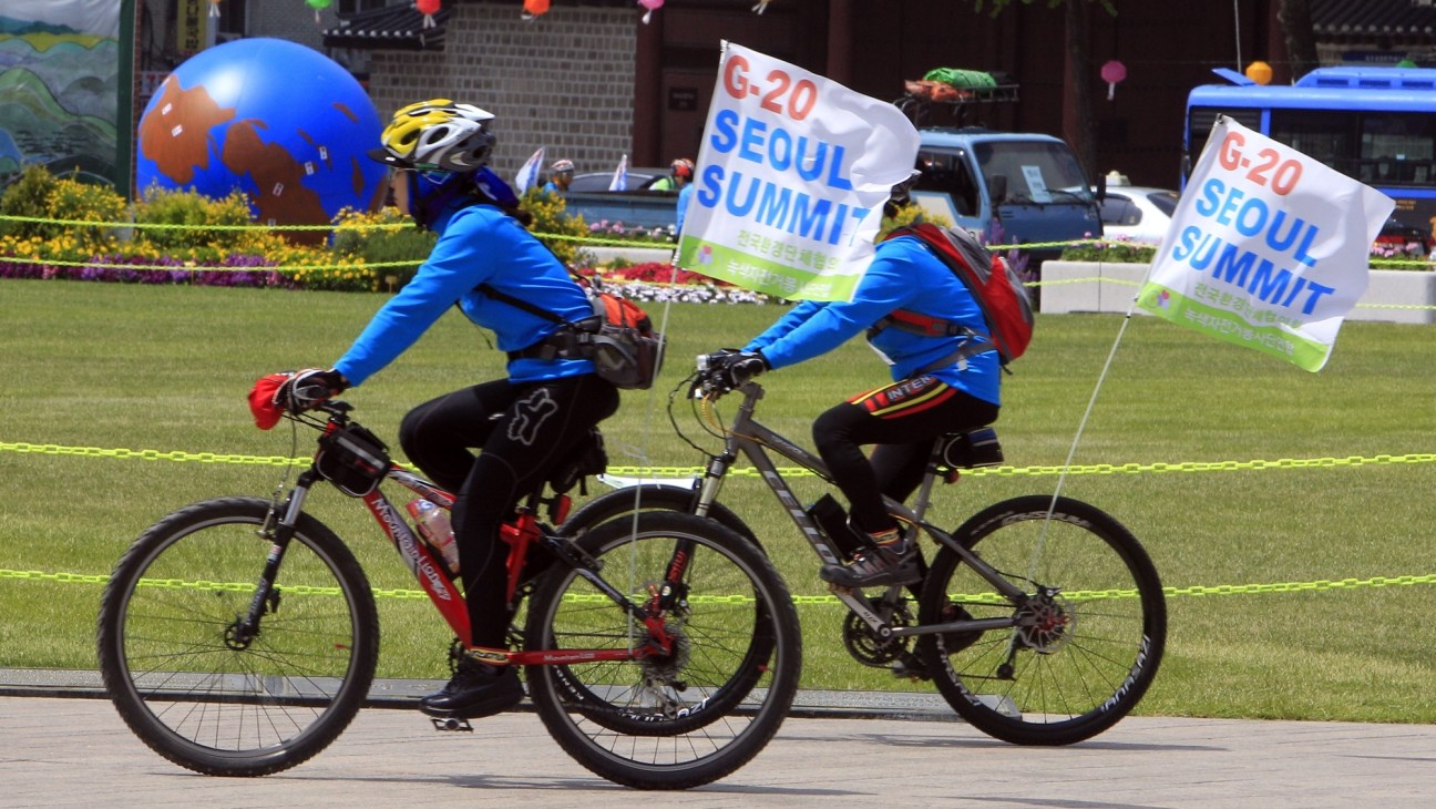 Two cyclists in blue jackets carry G20 Seoul Summit banners on bicycles during an outdoor event.
