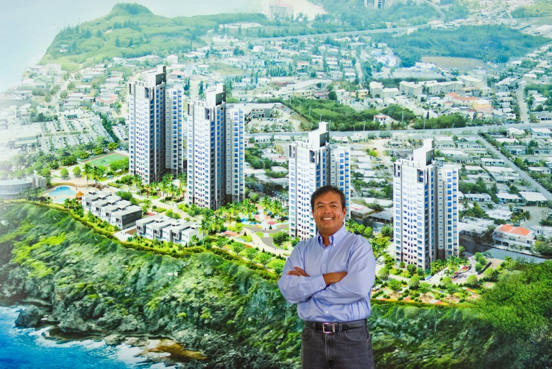 David Tydongco from Younex International stands with arms crossed before a rendering of the Emerald Oceanview Park residential towers on Guam's coast.