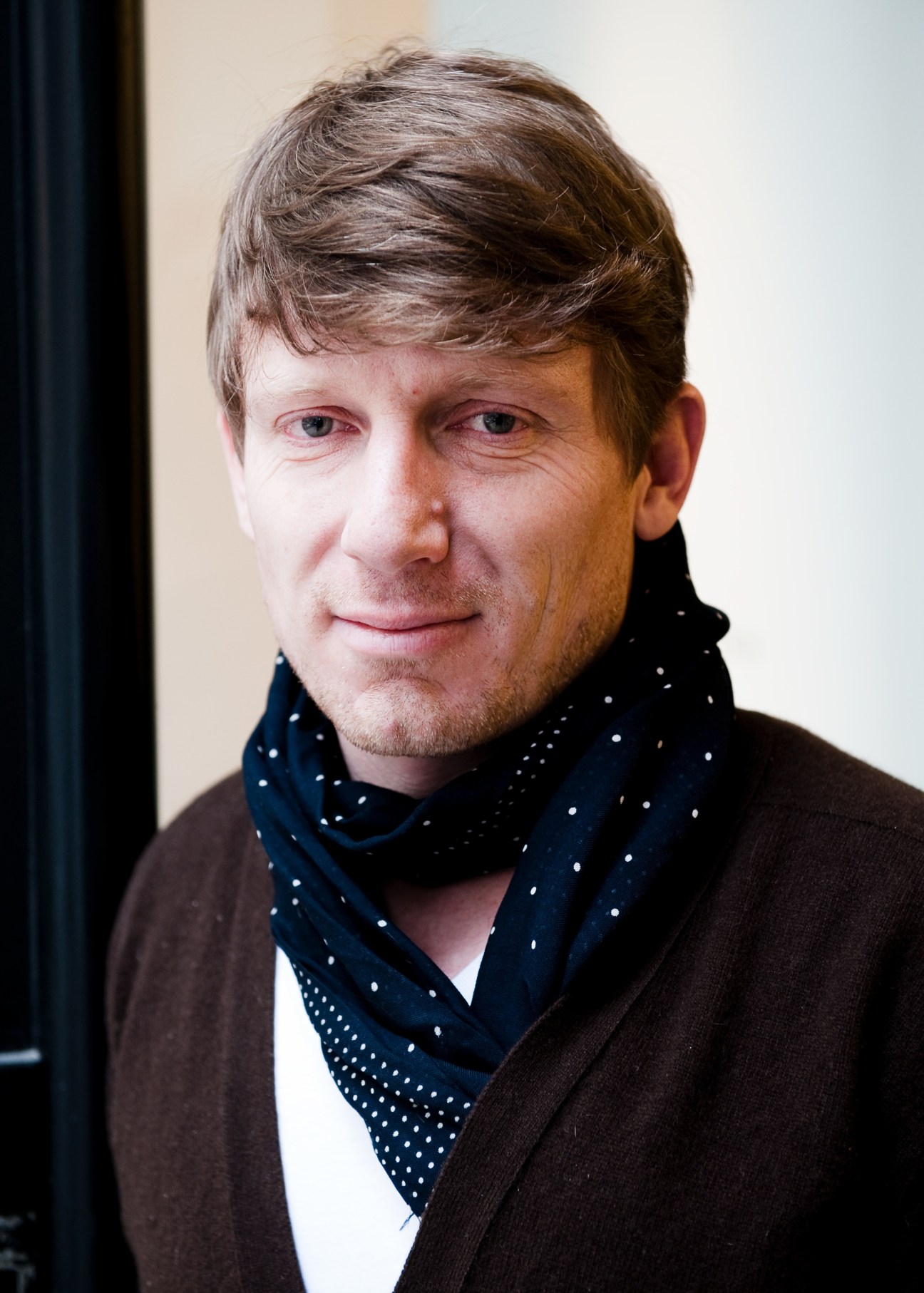 Portrait of Klaus Mühlbauer, co-owner of the Vienna hat-making firm, wearing a dark jacket and patterned scarf.