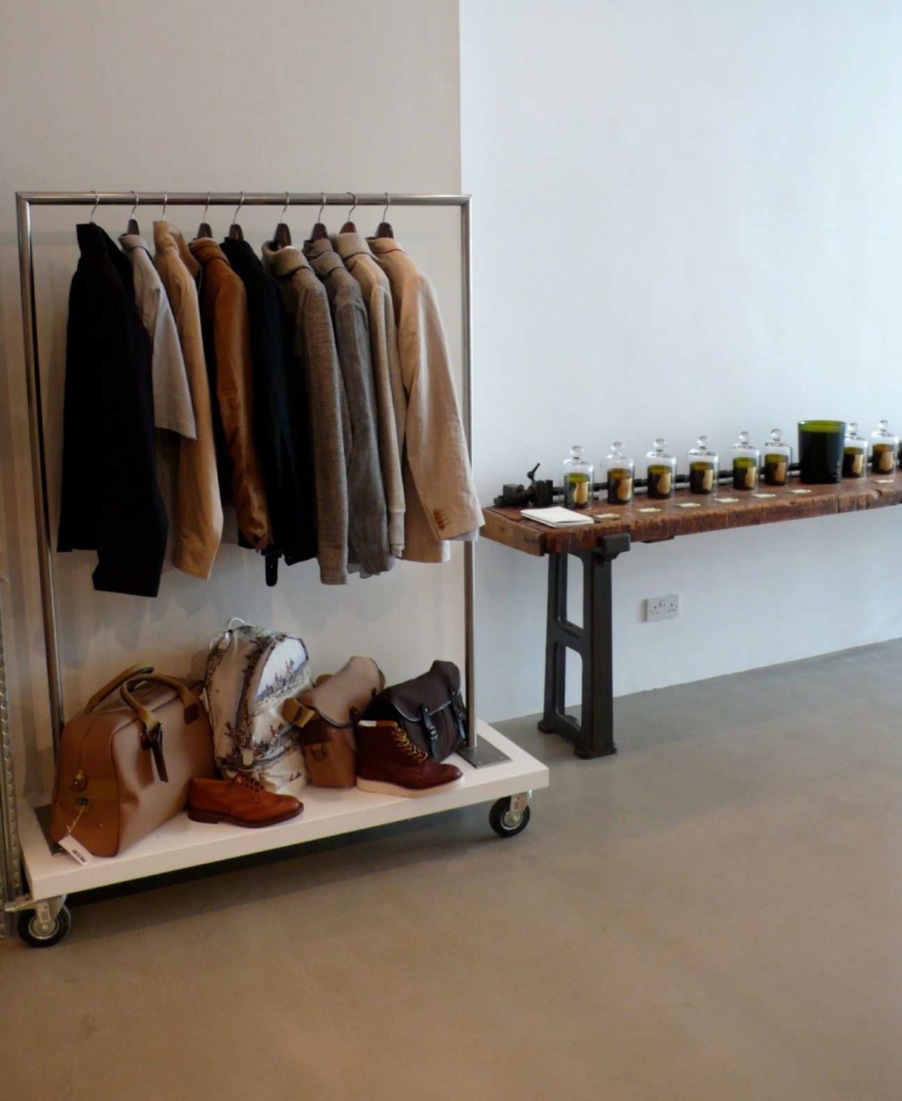 Present retail store interior with clothing rack displaying cardigans and shoes below, and scented candles on a wooden table.