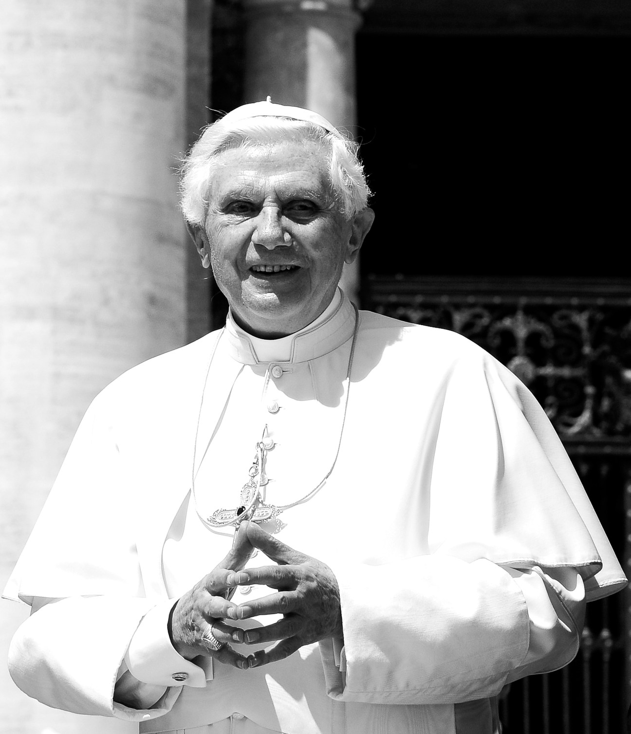 Pope Benedict XVI in white vestments and skull cap, holding a crucifix, photographed in black and white.