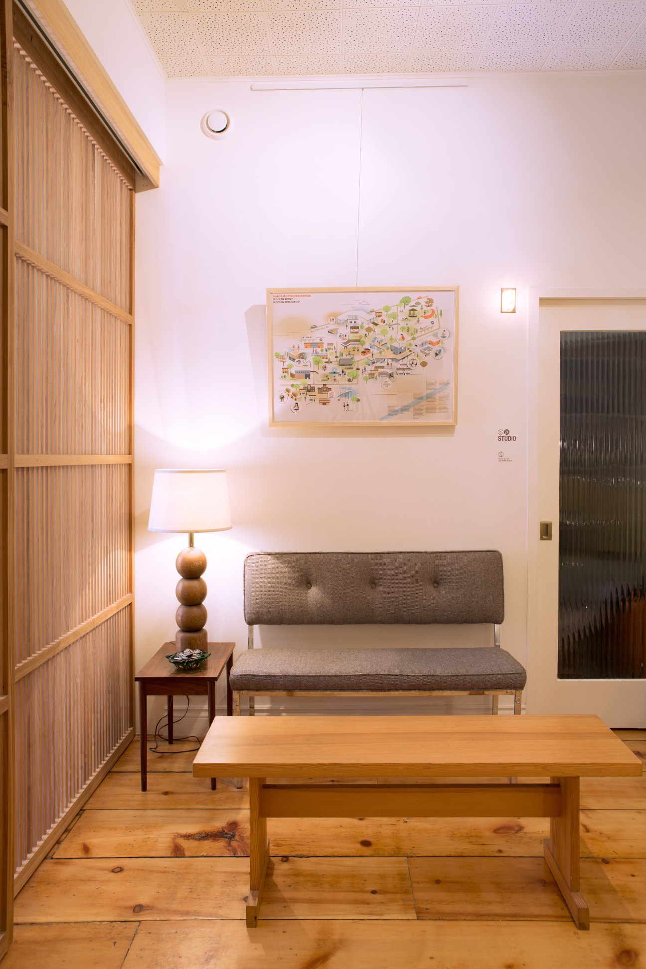 Waiting area with wooden table, gray upholstered bench, table lamp and framed map on white wall.