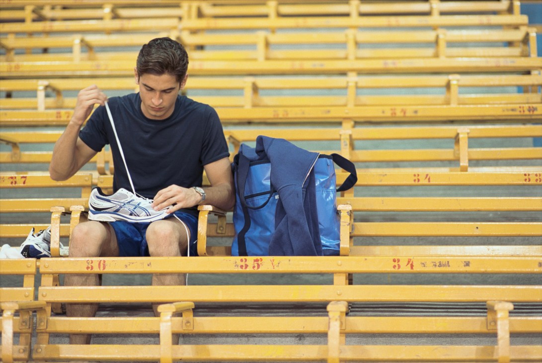 Model sitting on yellow bleachers wearing Dunhill t-shirt and Asics shorts, holding Asics shoes.