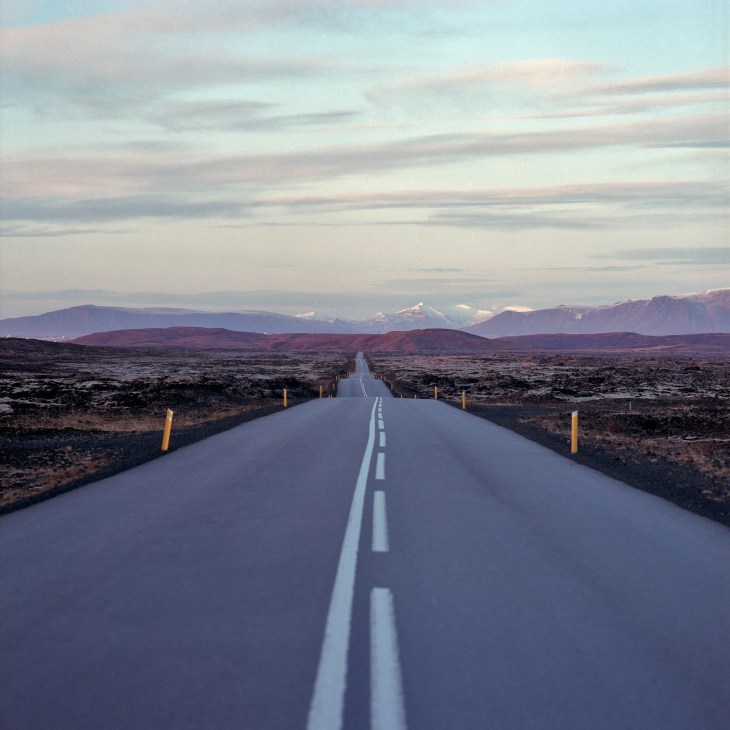 Straight road stretching through barren Icelandic lava landscape with snow-capped mountains in distance.