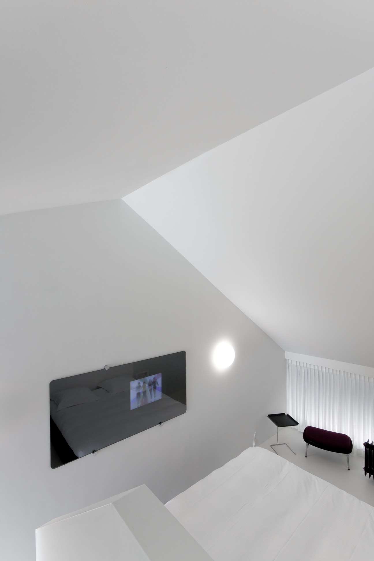 Minimalist hotel bedroom with angular white ceiling, embedded television in black wall panel, and modern furnishings.