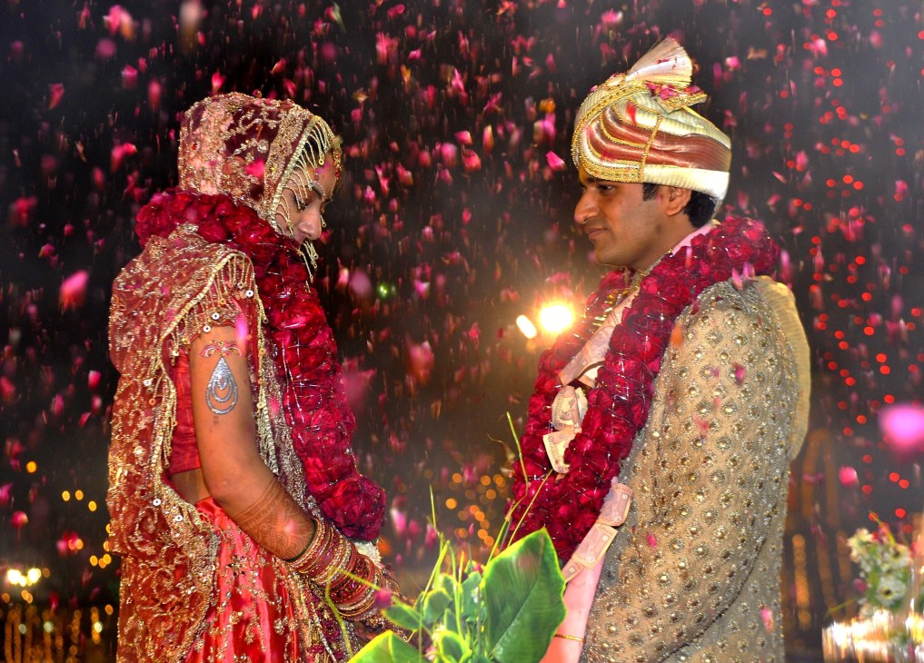 Couple in elaborate traditional Indian wedding attire with decorative flowers and lighting effects.