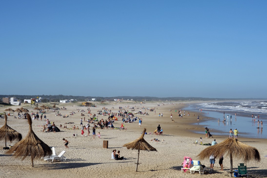 Wide sandy beach with visitors, thatched umbrellas, gentle waves, and coastal buildings in the distance under clear blue sky.