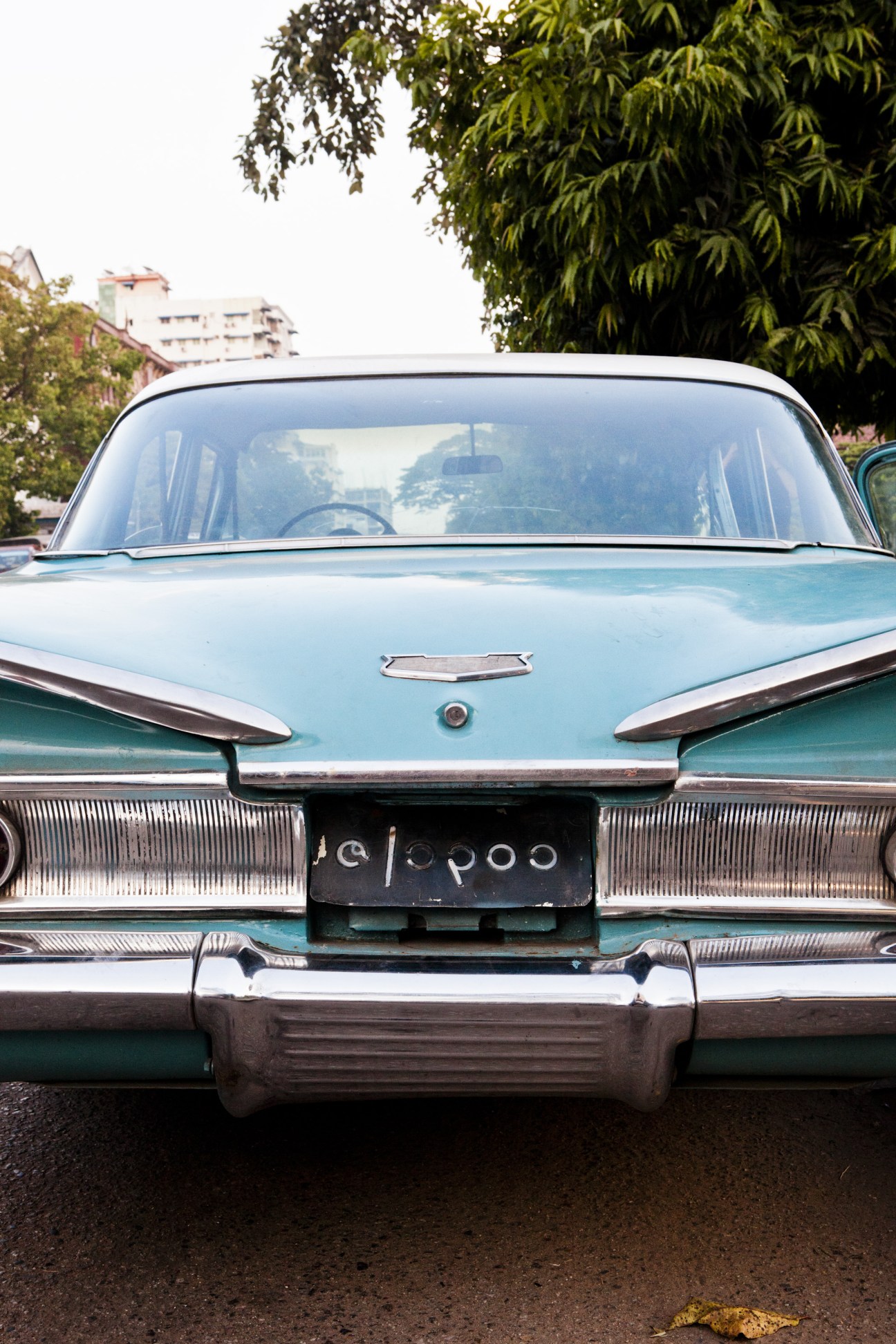 Front view of a turquoise vintage Cadillac parked on a Rangoon street with colonial buildings in the background.