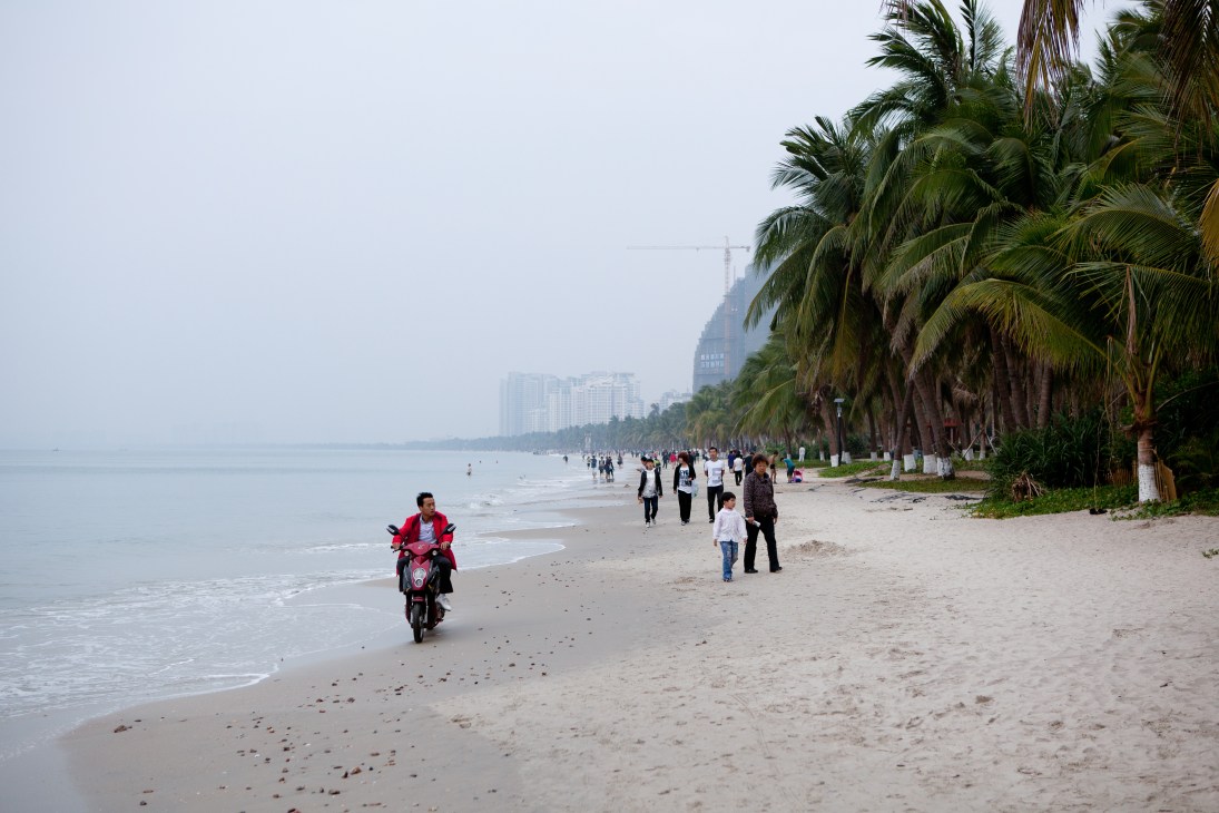A sandy beach in Sanya Bay with visitors, palm trees, and high-rise buildings visible in the misty distance.
