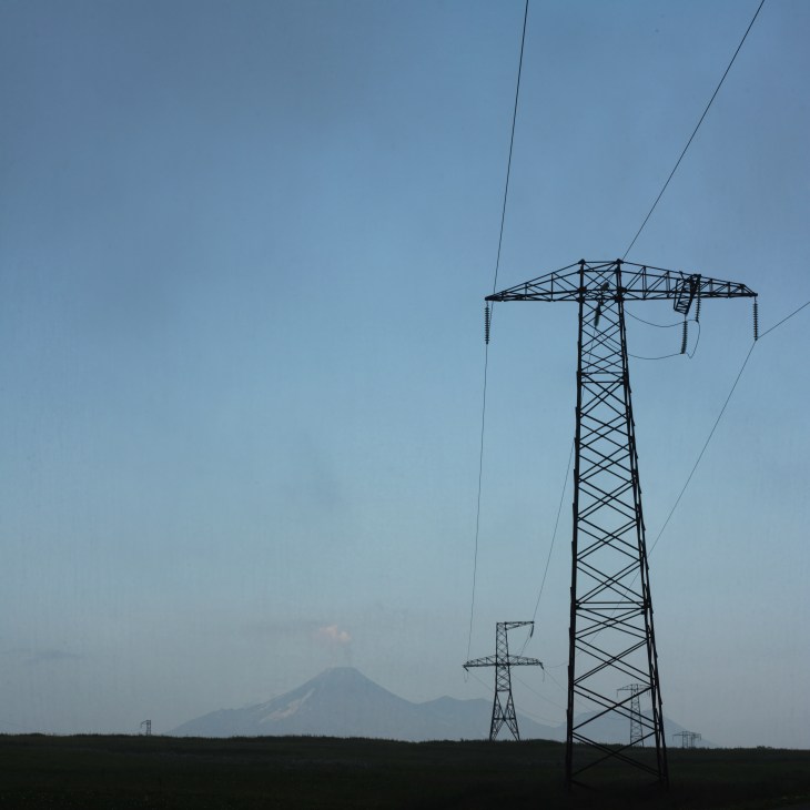 Electrical transmission pylons silhouetted against a misty landscape with the smoking Koryaksky volcano in the background.