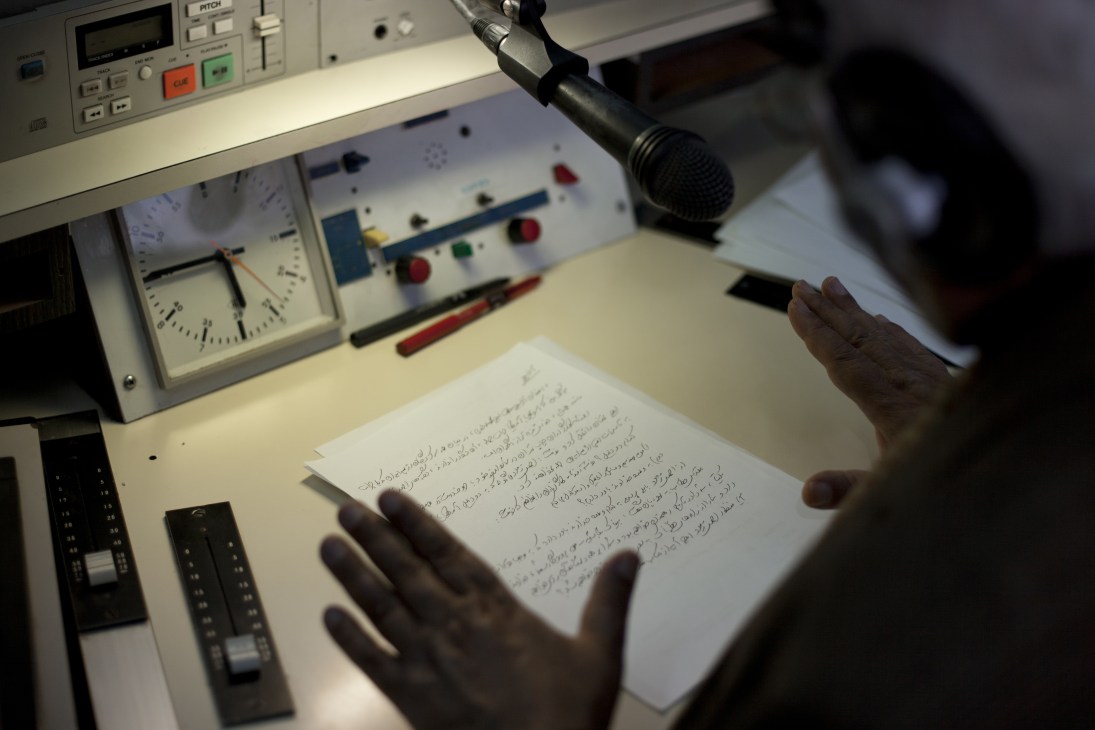 A broadcaster's hands holding a Farsi-written script at a radio studio desk with microphone and equipment.