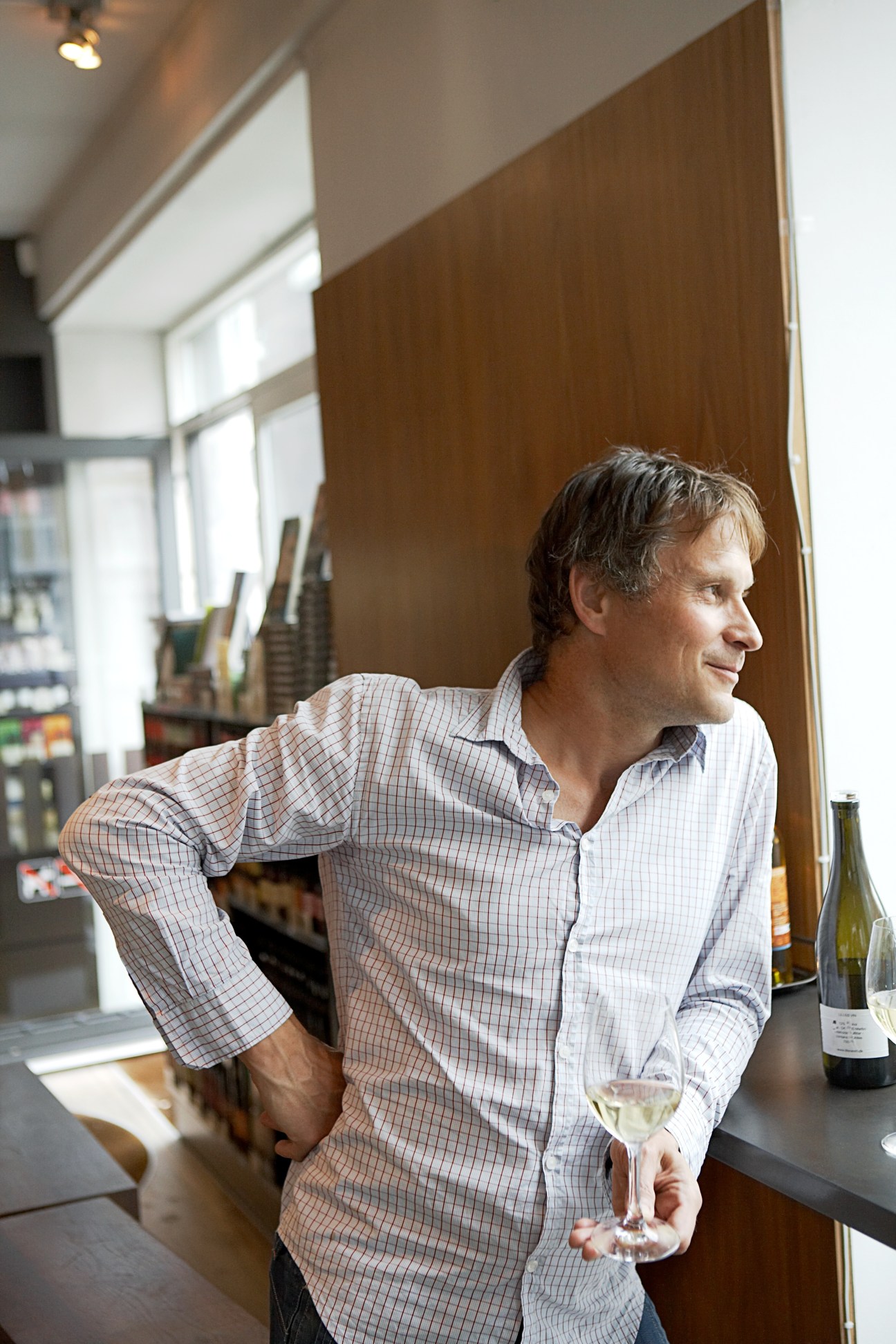 Claus Meyer stands in his kitchen holding a glass of white wine, wearing a light checkered shirt.
