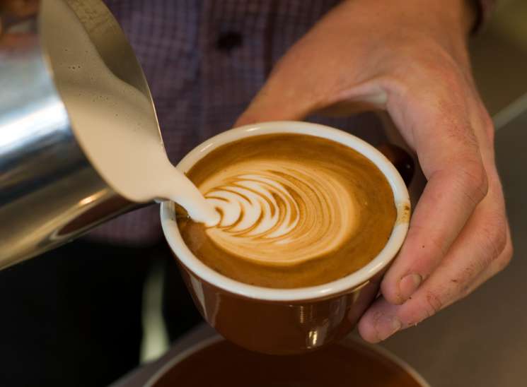 A flat white coffee with latte art held by a barista in a checkered shirt.