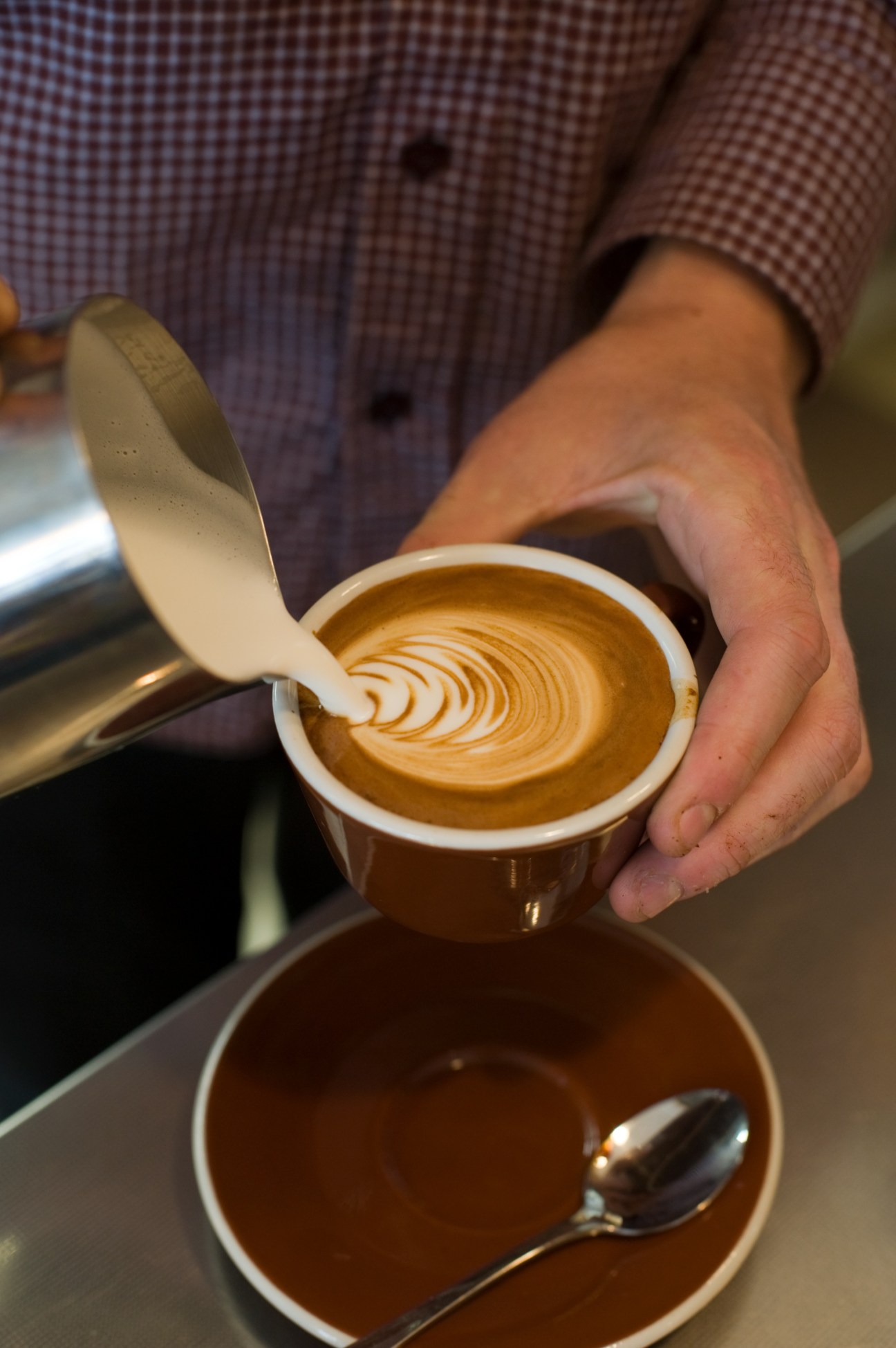 A flat white coffee with latte art held by a barista in a checkered shirt.