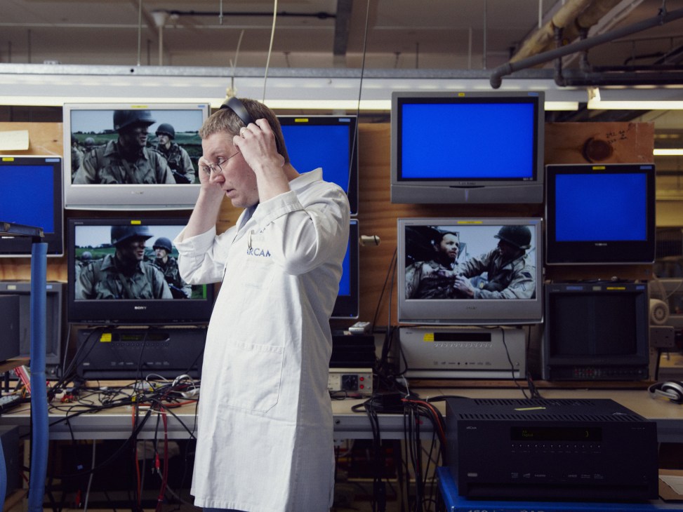 Arcam technician wearing white coat tests audio-video receivers surrounded by multiple monitors displaying video content.