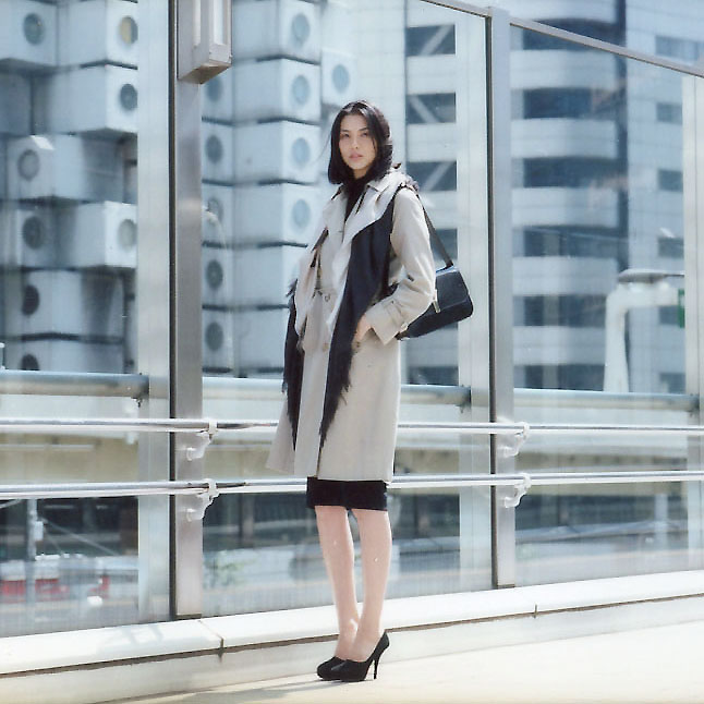 Woman wearing Aquascutum coat and Bottega Veneta dress poses against modern glass building backdrop.