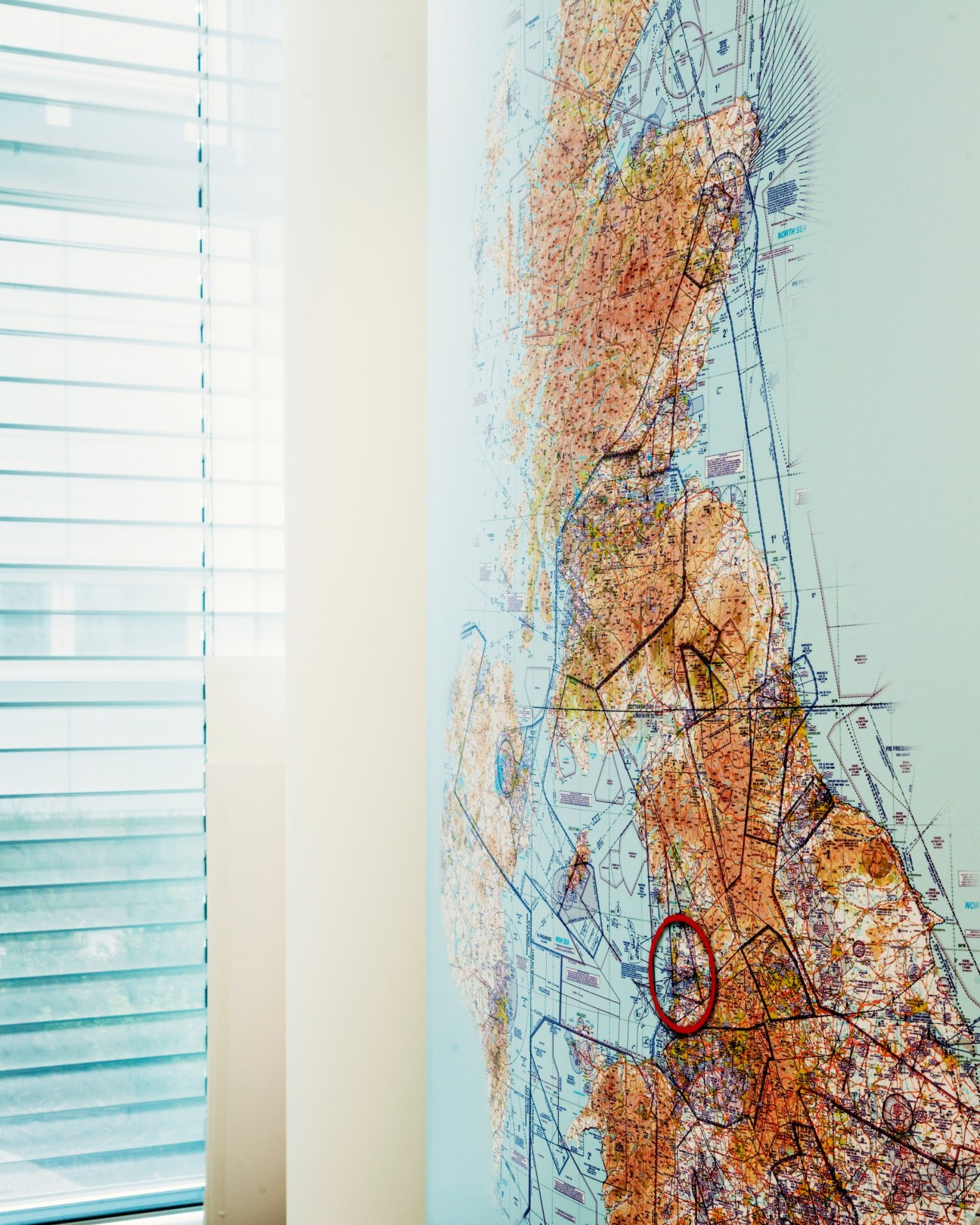 An air chart of the UK displayed on a wall next to a window with venetian blinds, showing terrain and flight paths marked with red circles.