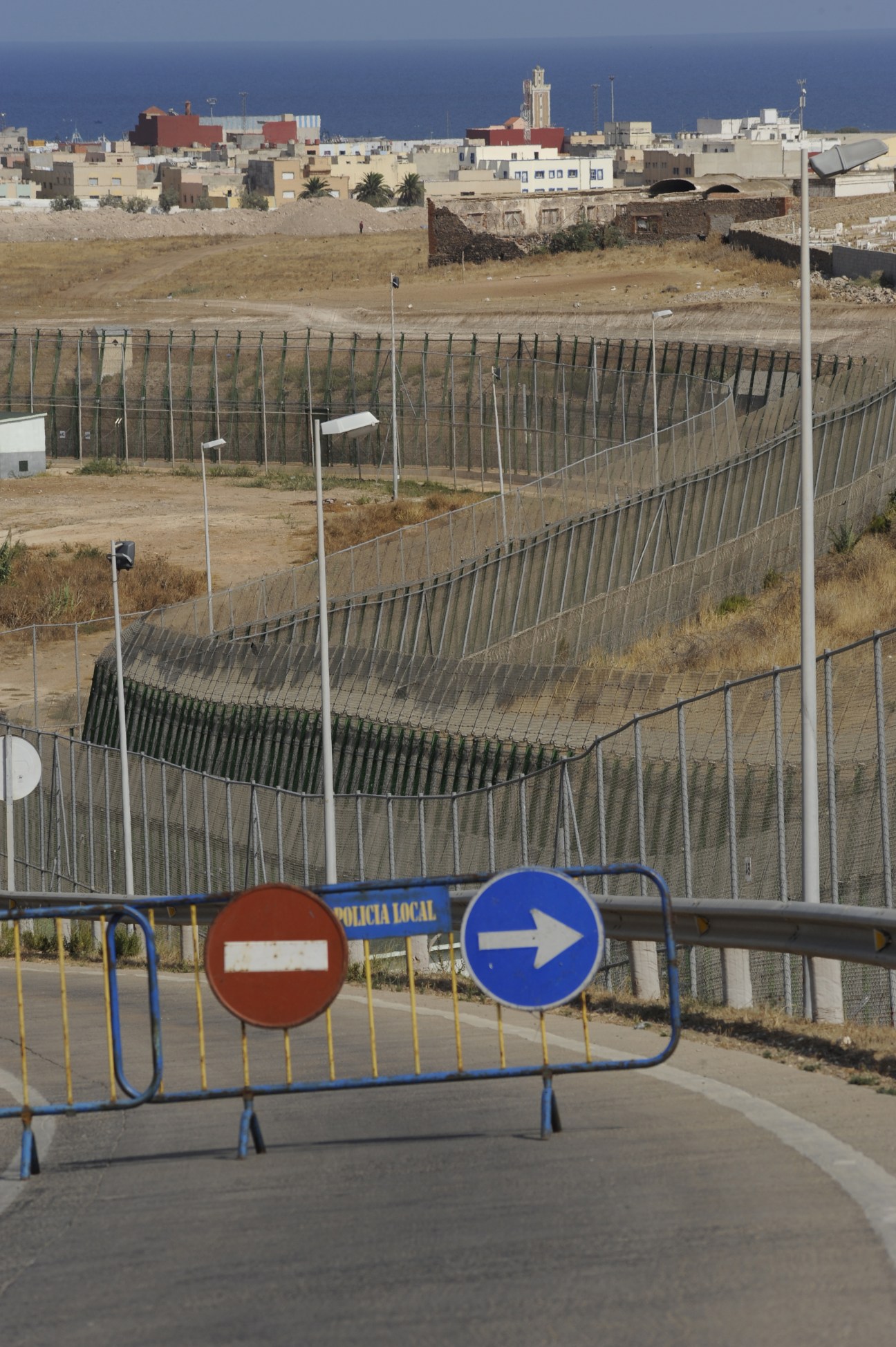Melilla's six-metre-high border fence with barbed wire stretching across dry terrain, with the city's buildings visible beyond.
