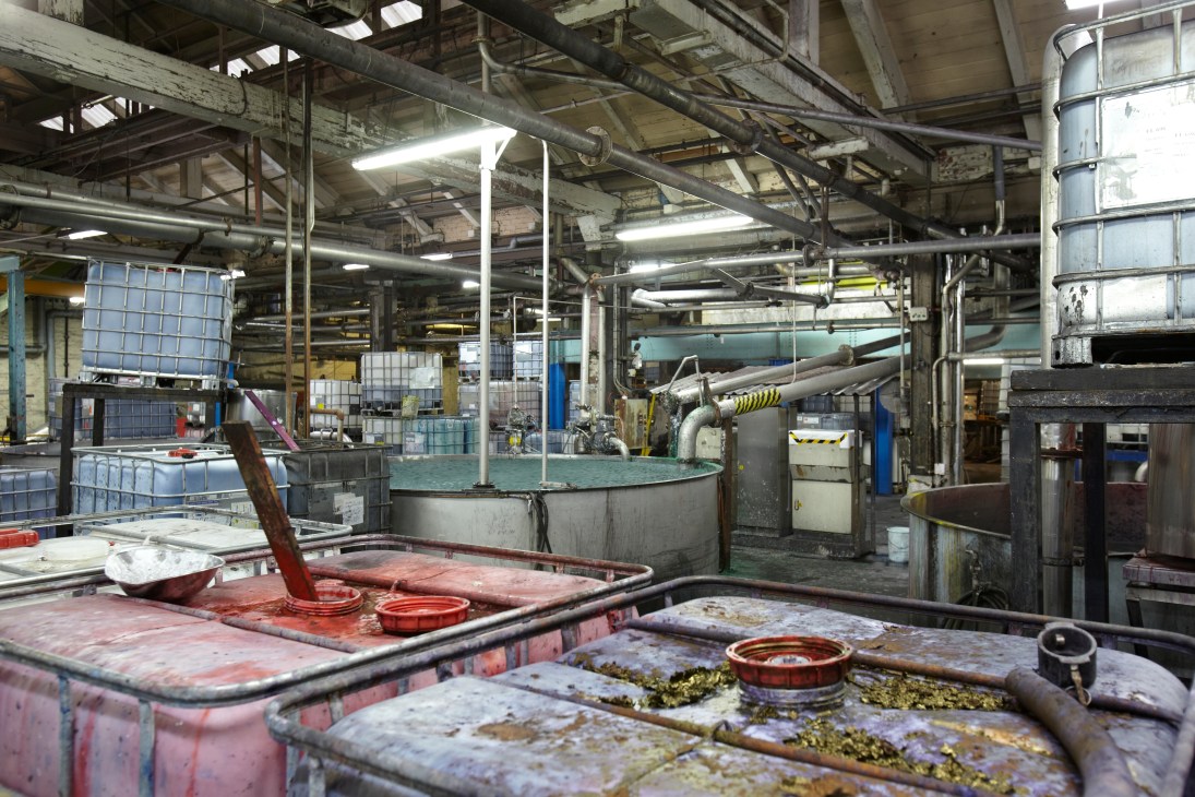 Industrial paper mill interior with large metal vats containing red and blue liquid dye mixtures for pulp blending.