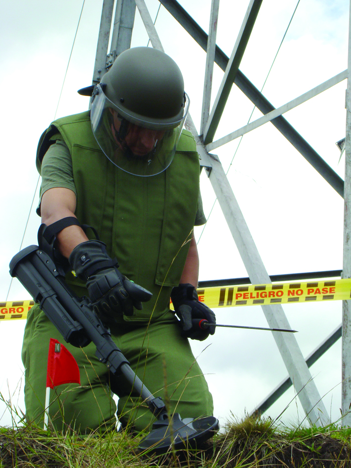 A deminer in protective green suit and helmet using specialized equipment to detect landmines in a field.