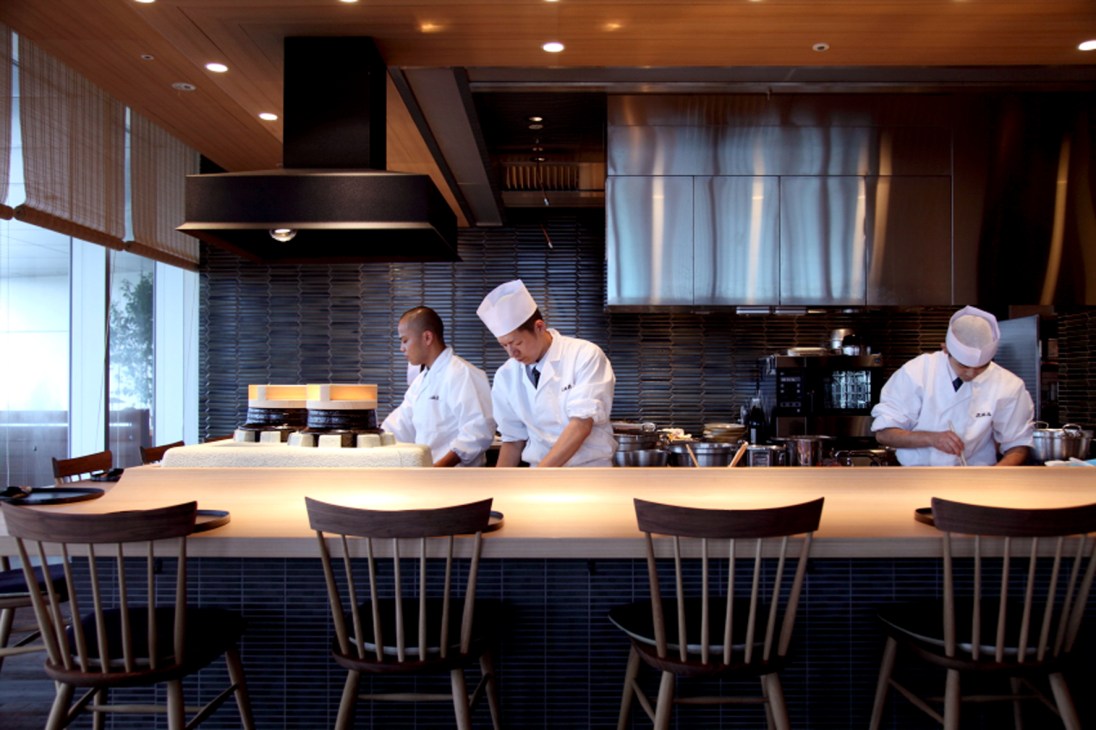Chefs in white uniforms and hats work behind a counter at a modern restaurant kitchen with dark tile backsplash.