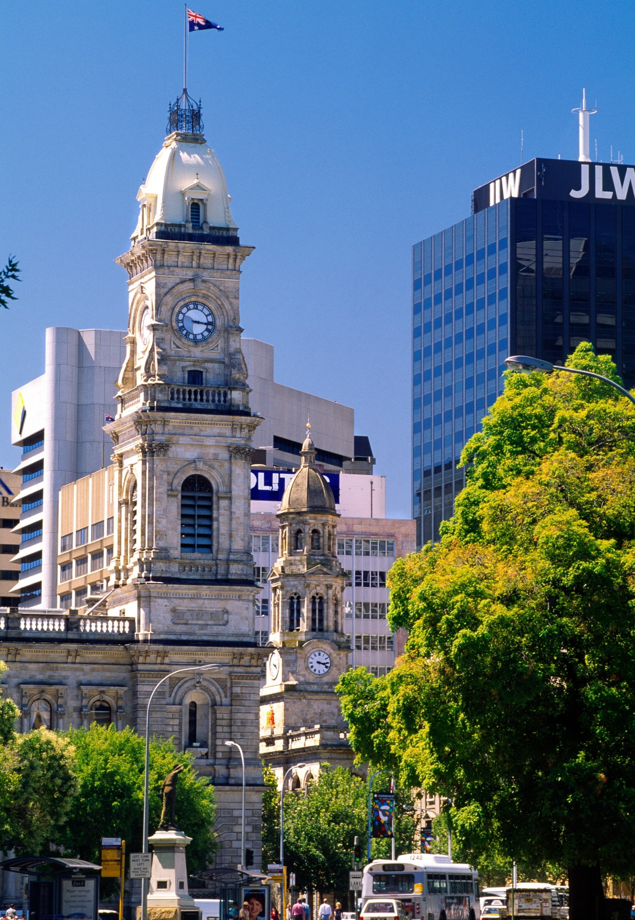 Melbourne streetscape with historic clock tower, modern buildings, and lush green trees.