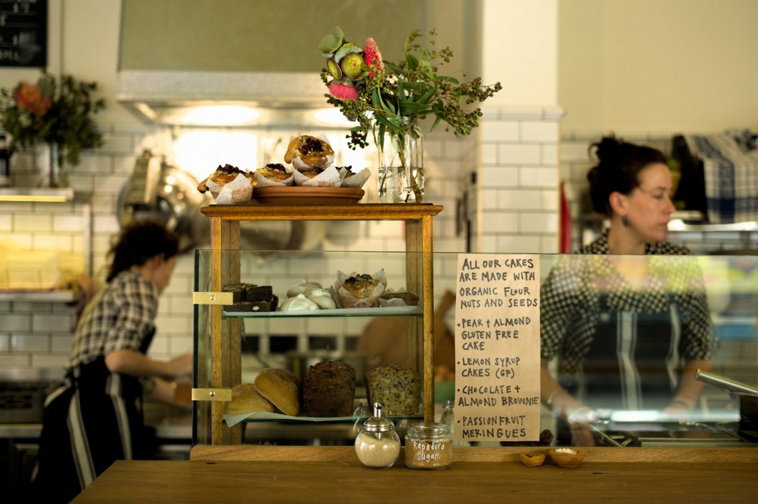 Display case at Cornersmith café with pastries, cakes, and fresh flowers on wooden shelf unit.
