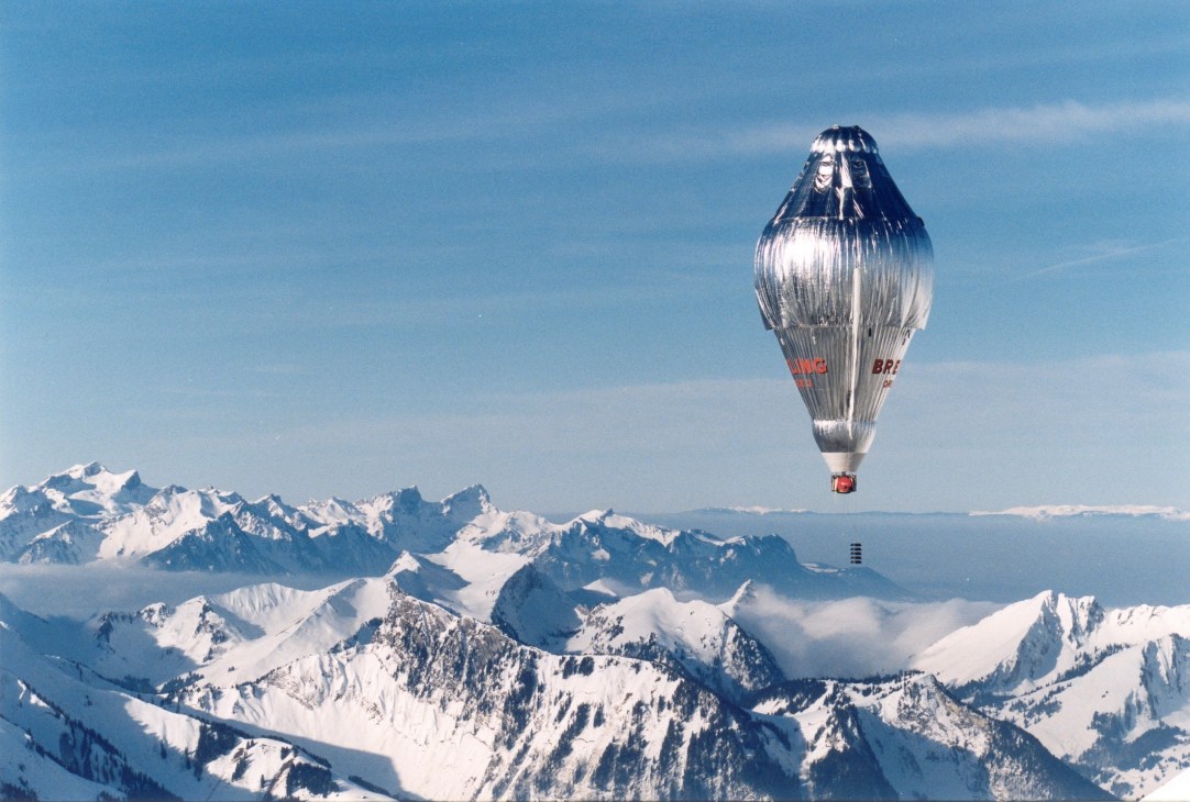 The Orbiter 3 hot-air balloon flying above snow-covered Alpine mountains under clear blue sky.