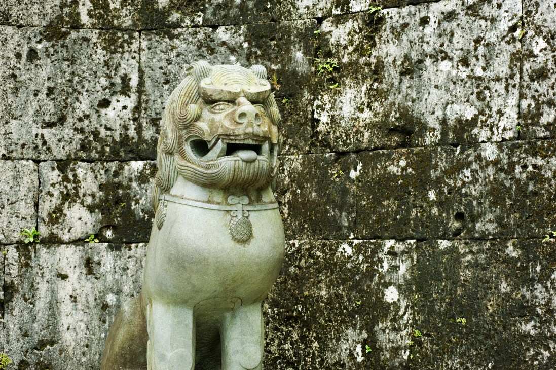 Stone shisa lion statue with open mouth against lichen-covered stone wall at Kankai gate.
