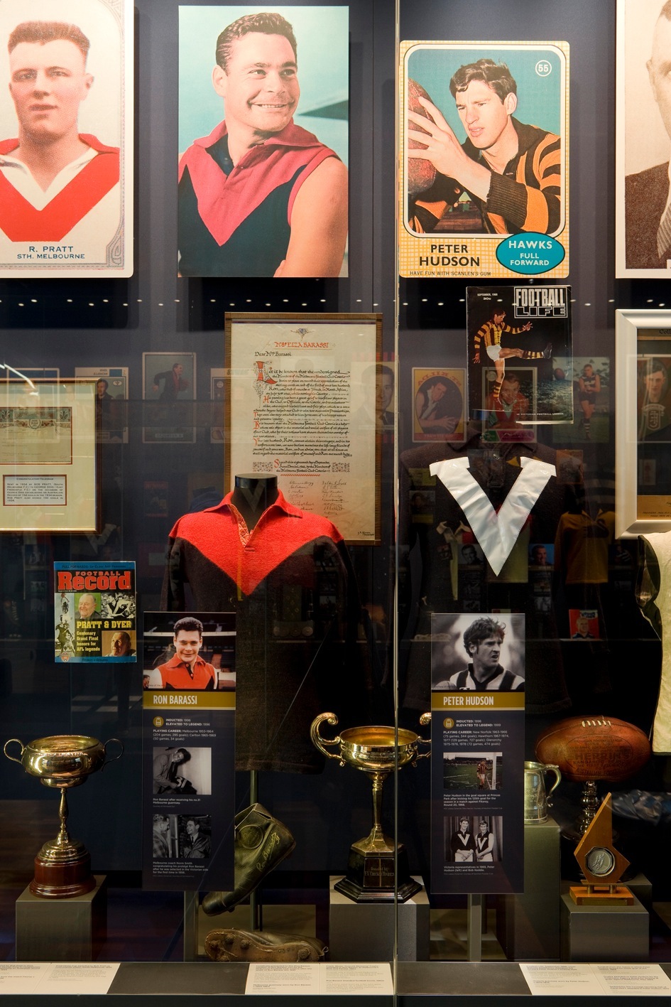 Museum display case showing Ron Barassi memorabilia including vintage football jerseys, trading cards, trophies and historical documents.