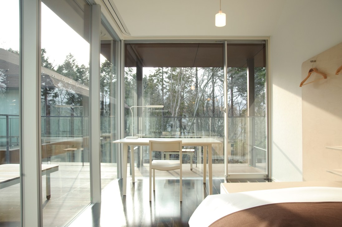 Minimalist bedroom with desk, wooden chair, and expansive glass doors overlooking forest.