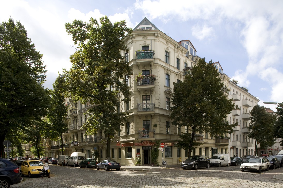 Pre-war Gründerzeit building with ornate stone facade, wrought iron balconies and tall trees lining a cobblestone street in Neukölln.
