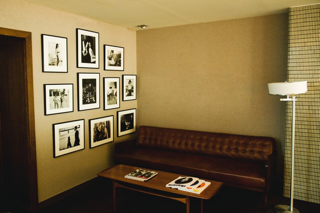 André Balazs Properties New York office meeting area with brown leather couch, framed photographs on mustard wall, and modern floor lamp.