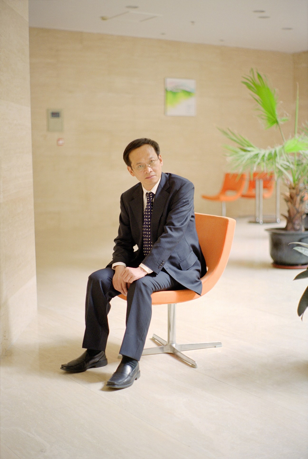 Man in business suit sitting on orange chair in modern office lobby with plant and signage