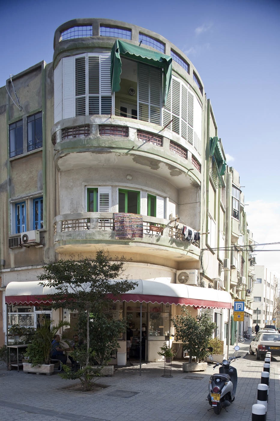 A curved Bauhaus building with green shutters and balconies, housing Miznon Avi restaurant with red awning at street level.