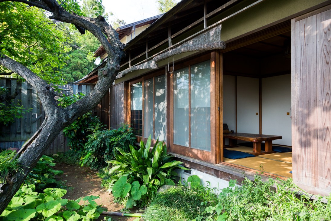 Traditional Japanese soba restaurant exterior with wooden architecture, shaded by a large tree, featuring sliding doors and a garden courtyard.