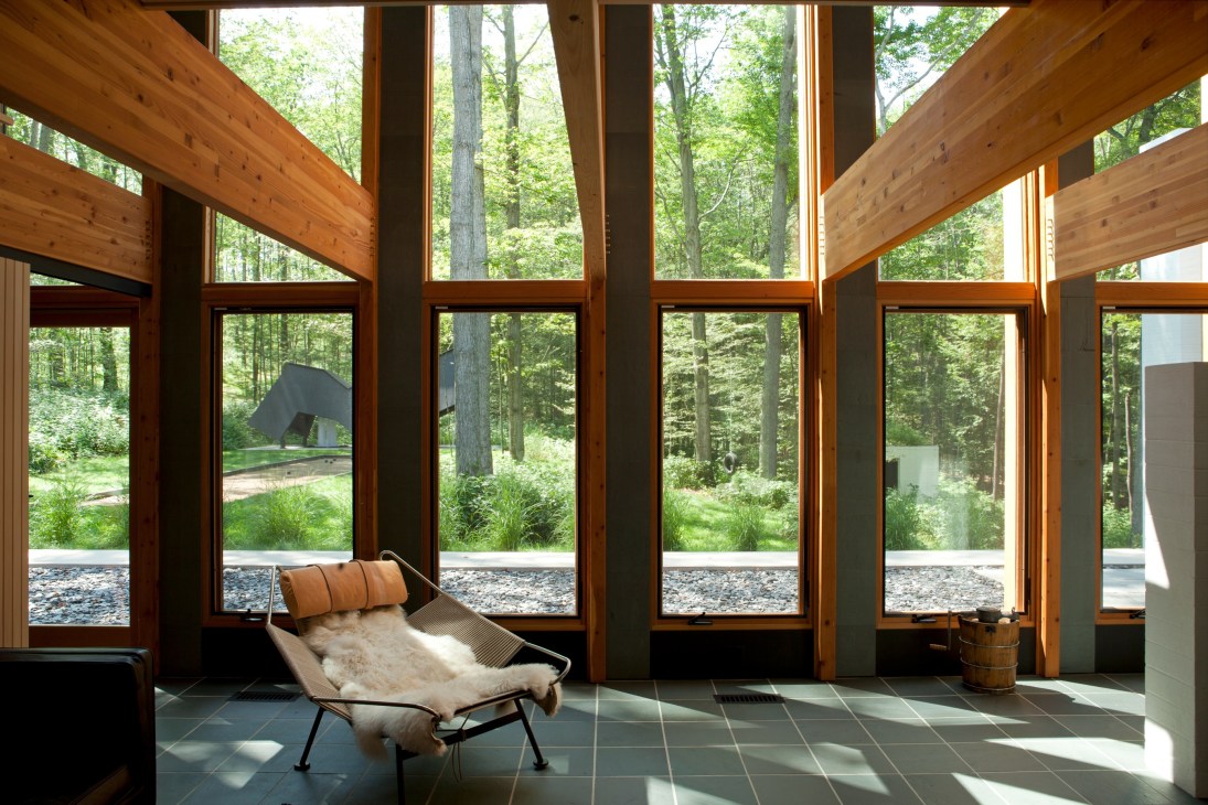 Modern interior with wooden beams and floor-to-ceiling windows overlooking a forest with tall trees.