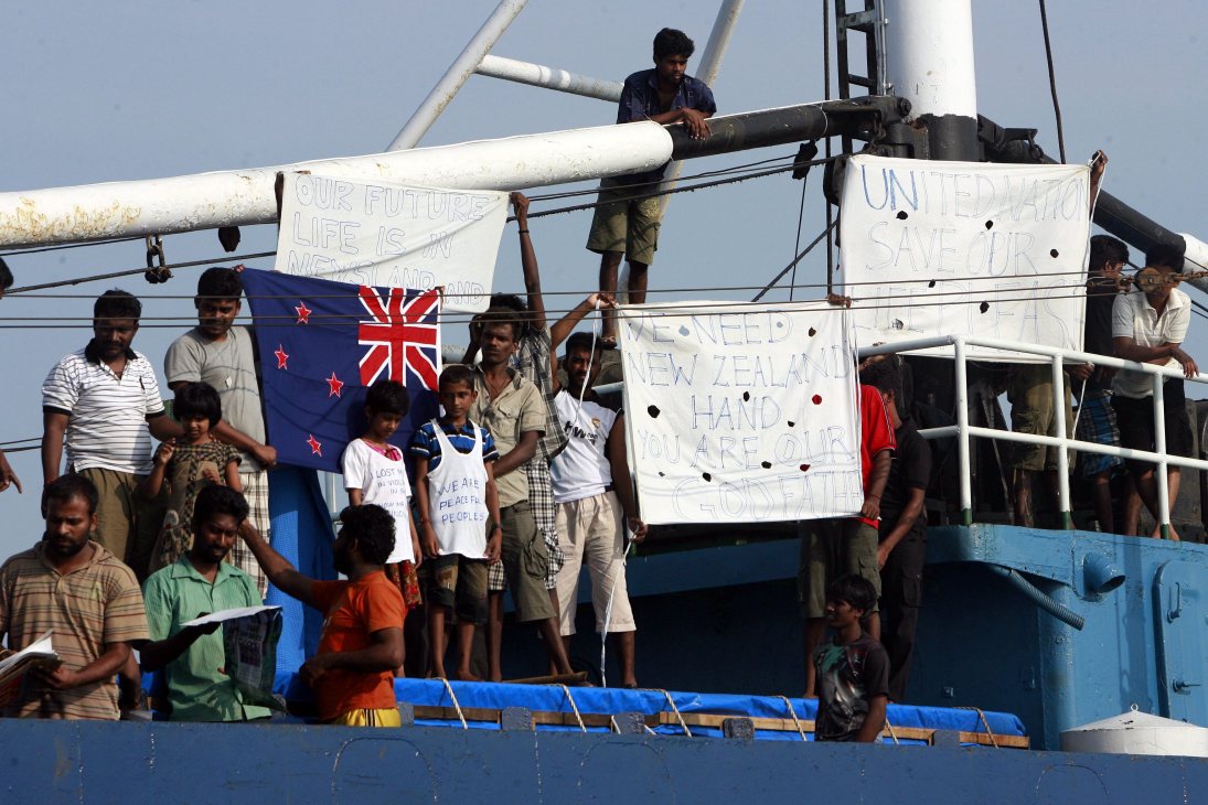 People on a ship displaying the New Zealand flag and handmade signs with messages about their future and need for help.
