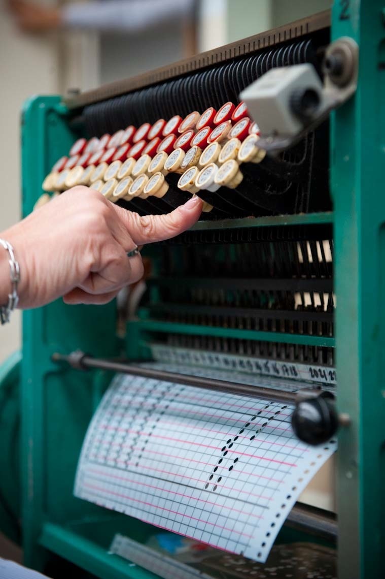 A hand holds leather threads inserted into a vintage loom with a punch card guide visible below.