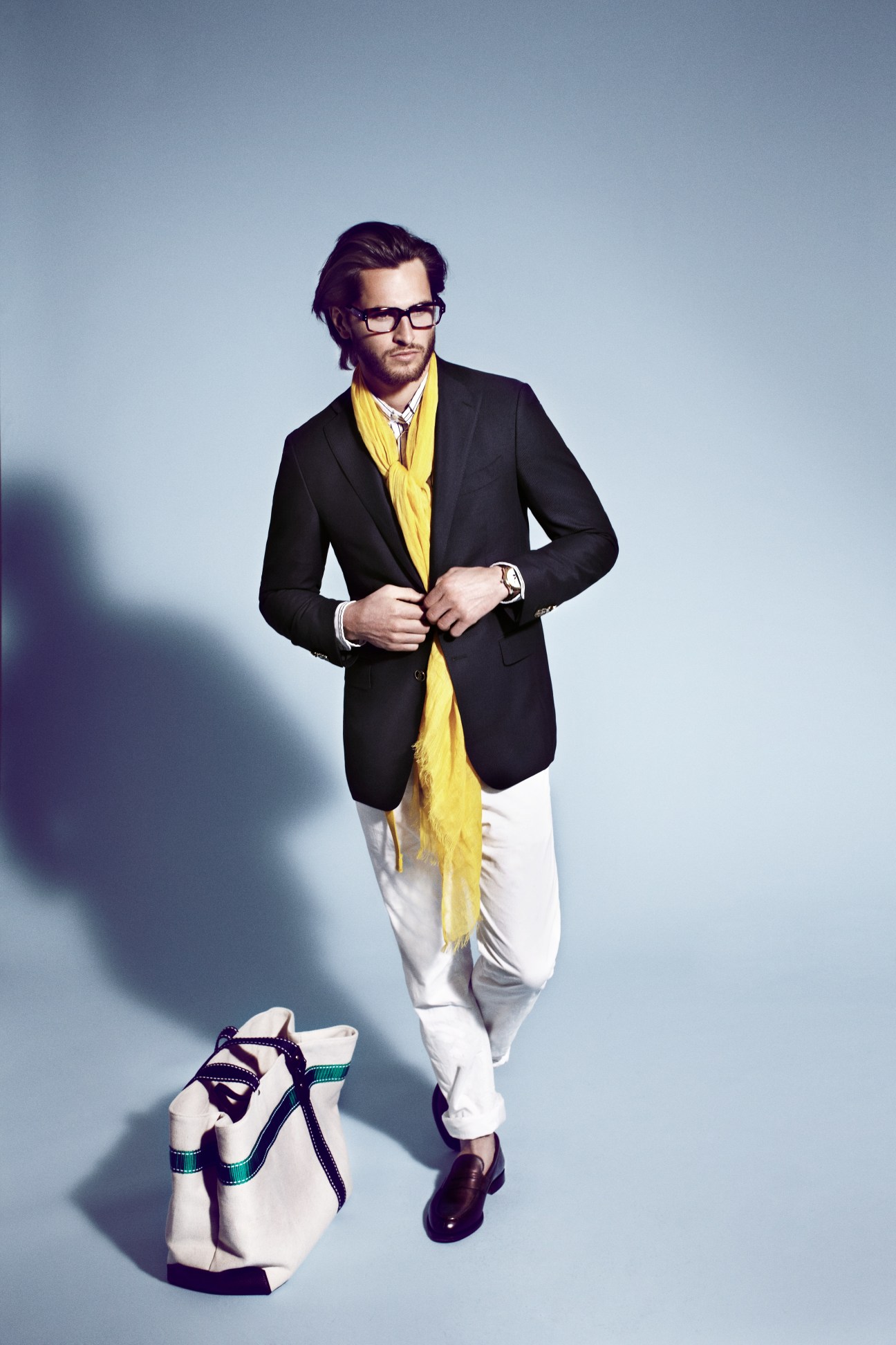 Male model wearing black Salvatore Ferragamo jacket, yellow scarf, white Bottega Veneta trousers, and Church's shoes against blue background.