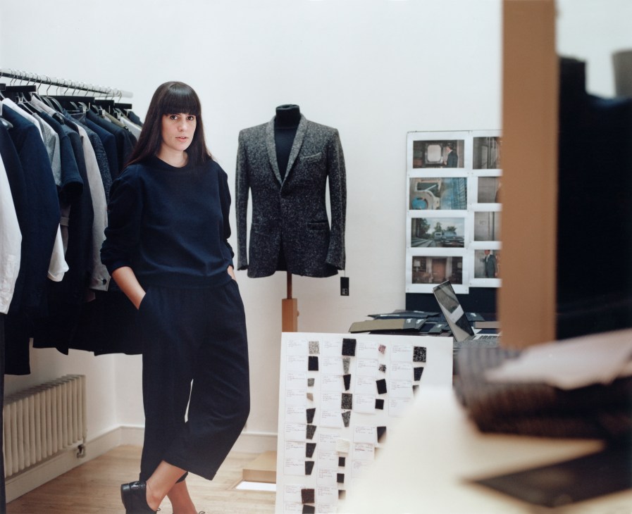 Paula Gerbase stands in her design studio surrounded by clothing samples, fabric swatches, and a mannequin wearing a gray blazer.
